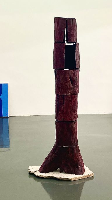 Abstrakte Skulptur aus dunkelrotem Material, auf einer hellen Basis stehend.